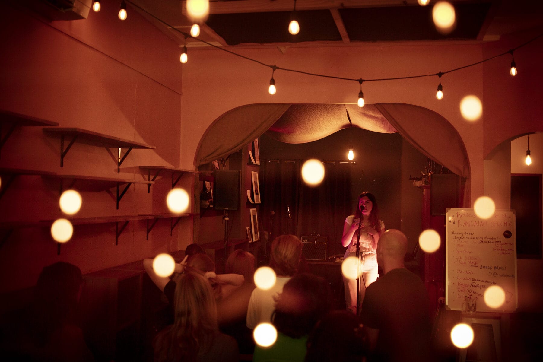 A warm-lit room of people watching a performer under festoon lights