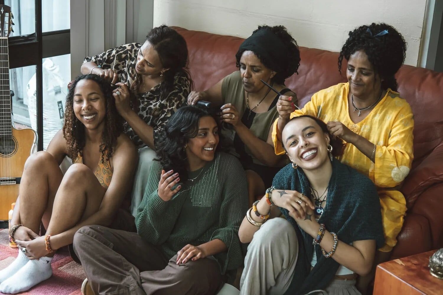 Young women laughing on the floor while their elders braid their hair on the couch behind them